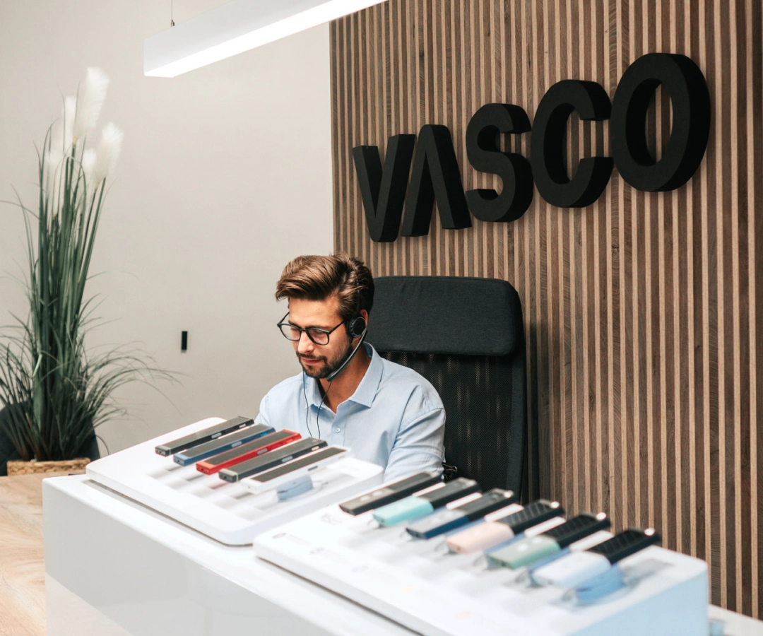 En man från kundtjänsten sitter vid ett skrivbord och visar upp flera Vasco Translator-enheter i olika färger, med en stor