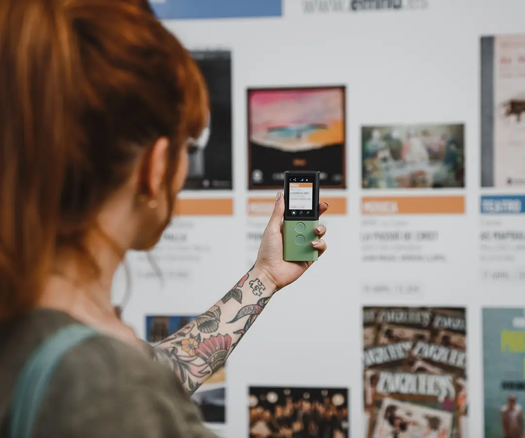 En person med rött hår och en blommig tatuering på armen håller upp en grön digital enhet och pekar den mot en anslagstavla med olika affischer och information om evenemanget.
