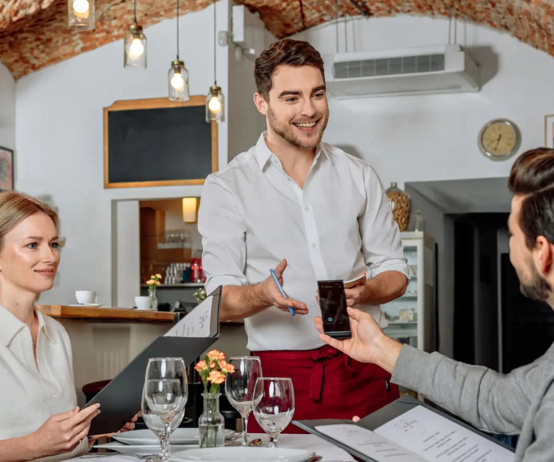En leende servitör står vid ett restaurangbord, medan en sittande manlig kund håller en översättare i handen. En kvinnlig kund sitter i närheten och håller i en meny. Bordet är dukat med glas, tallrikar och en liten blomvas.