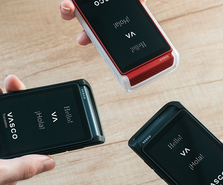 Tre personer som håller i Vasco Translator Q1-enheter samlas; på skärmarna visas startskärmen med Vasco-logotypen och orden ”Hello!” och ”¡Hola!”. Enheterna presenteras i silikonfodral i tre färger: Solid Black, Dark Caramel och Milky White.