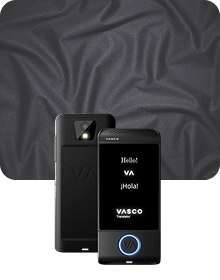 En produktbild som visar en svart Vasco Translator-enhet från två vinklar mot en mörk bakgrund av texturerat tyg. Enheten till vänster visar baksidan, där kameralinsen med blixt och det präglade ”VA”-logotypen framhävs. Enheten till höger visar framsidan med upplyst skärm, där hälsningarna ”Hello!”, ”VA”, ”¡Hola!” och ”VASCO Translator”-logotypen visas. En rund knapp längst ned på enhetens framsida är kantad med ett lysande blått ljus. Bakgrunden består av draperat, kolgrått tyg med en subtil nätliknande struktur.