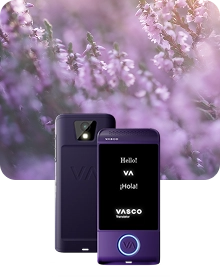 En produktbild som visar en lila Vasco Translator-enhet från två vinklar mot en mörk bakgrund av texturerat tyg. Enheten till vänster visar baksidan, där kameralinsen med blixt och det präglade ”VA”-logotypen framhävs. Enheten till höger visar framsidan med upplyst skärm, där hälsningarna ”Hello!”, ”VA”, ”¡Hola!” och logotypen ”VASCO Translator” visas. En cirkulär knapp längst ned på enhetens framsida är kantad med ett lysande blått ljus. Kompositionen är placerad mot en drömlik, suddig bakgrund av lila ljungblommor badade i mjukt solljus.