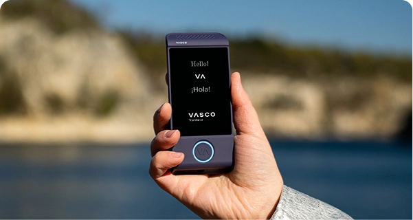 En närbild av en hand som håller en lila elektronisk översättningsenhet för baskiska mot en ljus utomhusbakgrund. På skärmen visas hälsningarna ”Hello!”, ”¡Hola!” och logotypen för ”VASCO Translator”. I bakgrunden syns en bild med mjuk fokusering av ett soligt kustlandskap med blått vatten och ljusa klippor under en klar himmel. Personen som håller enheten bär en ljusgrå eller silverfärgad jackärm med struktur. 