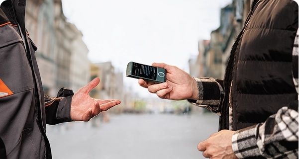 En person klädd i en svart, quiltad väst över en rutig skjorta håller en turkosfärgad elektronisk översättare av märket Vasco horisontellt riktad mot en annan person på en gata i staden. Den andra personen, klädd i en grå och orange arbetsjacka, sträcker fram handen som om de just pratar med varandra. På enhetens skärm visas ett översättningsgränssnitt med text. I bakgrunden syns en vidsträckt, suddig vy av en gata i europeisk stil med höga byggnader under en ljus, molnig himmel. 