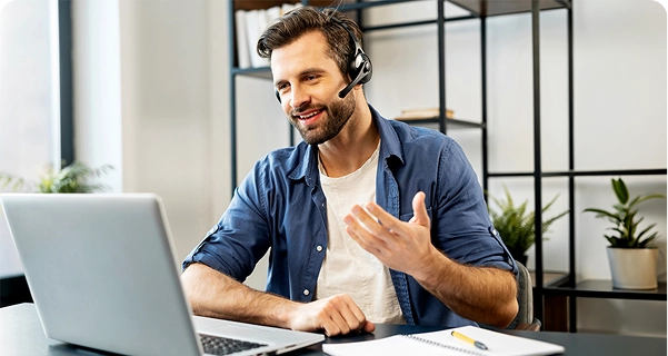 En vänligt utseende man med skägg och mörkt hår sitter vid ett skrivbord och bär ett professionellt headset med mikrofon. Han ler och gestikulerar med ena handen medan han tittar på en bärbar datorskärm; det ser ut som om han är mitt i ett videosamtal eller ger teknisk support. Han är klädd i en blå skjorta över en vit t-shirt. På skrivbordet framför honom ligger en bärbar dator, en anteckningsbok och en gul penna. Bakgrunden är ett ljust, modernt kontor med en minimalistisk svart metallhylla och gröna krukväxter. 