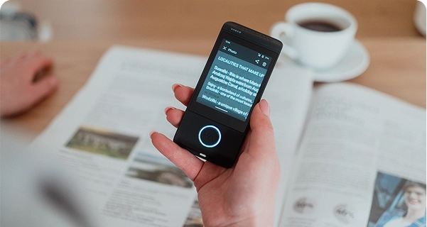 En bild tagen uppifrån som visar en persons hand som håller en svart elektronisk översättare av märket Vasco över en uppslagen tidning på ett träbord. Enhetens skärm är i ”Foto”-läge och visar en inzoomad, översatt engelsk text om lokala sevärdheter. I bakgrunden syns en vit kaffekopp och tidningens kanter, vilket skapar en avslappnad miljö för läsning och översättning. 